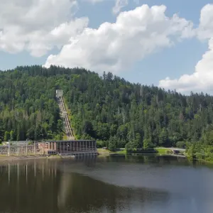 Eine Außenaufnahme eines Wasserkraftwerks an einem See. Im Hintergrund erhebt sich ein bewaldeter Hügel mit einer langen, steilen Rohrleitung, die von einem Gebäude auf der Hügelkuppe hinabführt. Das Kraftwerk befindet sich am Ufer des Sees und besteht aus 