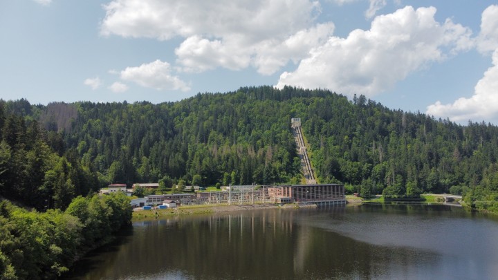 Eine Außenaufnahme eines Wasserkraftwerks an einem See. Im Hintergrund erhebt sich ein bewaldeter Hügel mit einer langen, steilen Rohrleitung, die von einem Gebäude auf der Hügelkuppe hinabführt. Das Kraftwerk befindet sich am Ufer des Sees und besteht aus 