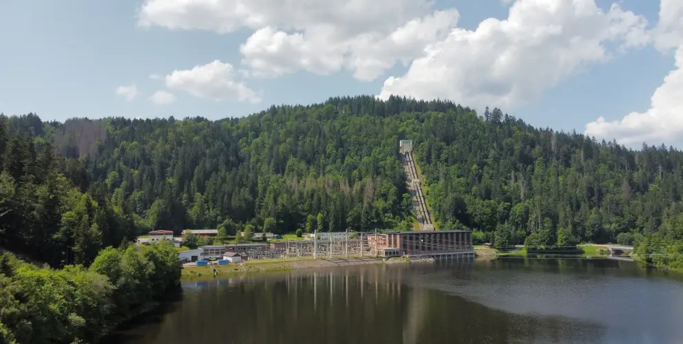 Eine Außenaufnahme eines Wasserkraftwerks an einem See. Im Hintergrund erhebt sich ein bewaldeter Hügel mit einer langen, steilen Rohrleitung, die von einem Gebäude auf der Hügelkuppe hinabführt. Das Kraftwerk befindet sich am Ufer des Sees und besteht aus 