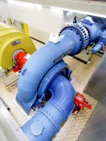 Ein Blick von oben auf eine große, blaue Wasserturbine in einer Industriehalle. Die Turbine ist mit einem gelben Generator verbunden, der ein rotes Bauteil an der Vorderseite hat. Der Boden ist mit kleinen Kacheln bedeckt, die Wände sind hell. Rohre und Leitungen verlaufen durch den Raum.