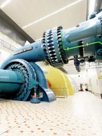 Eine industrielle Maschinenhalle mit einer großen blauen Wasserturbine von „Escher Wyss“. Die Turbine ist mit einem massiven Rohr verbunden, das an die Decke führt. Im Vordergrund ist ein rotes Steuerungselement mit verschiedenen Ventilen und Anzeigen zu sehen. Der Boden ist gefliest, und die Halle ist hell beleuchtet.
