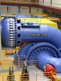 Eine große, blaue Wasserturbine der Marke „Escher Wyss“ in einer Industriehalle. Die Turbine hat massive Rohrleitungen und eine auffällige Biegung. Im Vordergrund sind Geländer und ein rotes Bauteil zu sehen. Im Hintergrund befinden sich weitere Maschinen, Metallgestelle und eine erhöhte Laufplattform.