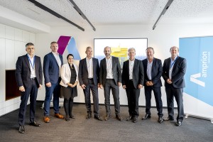 Acht Personen, darunter eine Frau, stehen in einem modernen Konferenzraum und posieren für ein Gruppenfoto. Alle tragen Business-Kleidung und wirken professionell. Der Hintergrund zeigt ein großes Logo in Violett und Blau, einen Bildschirm mit einer Offshore-Plattform sowie ein blaues Banner mit „Amprion verbindet“. Der Raum ist modern und gut beleuchtet.