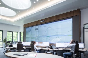 Das Bild zeigt einen modernen Kontrollraum mit mehreren Arbeitsplätzen, an denen zwei Personen arbeiten. Eine große Videowand dominiert die Wand und zeigt Diagramme und Datenanalysen. Vor der Wand stehen mehrere Schreibtische mit mehreren Monitoren, Laptops und Telefonen. Eine Person sitzt links und tippt auf der Tastatur, während die andere rechts telefoniert. Der Raum ist hell beleuchtet, hat eine moderne Decke mit runden Leuchtelementen und große Fenster, durch die grüne Bäume sichtbar sind. Die Atmosphäre wirkt professionell und technologisch fortschrittlich.