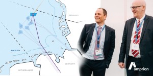 Zwei Männer in Anzügen mit blauen Konferenz-Badges stehen lachend vor einem großformatigen Kartenausschnitt der Nordsee und angrenzender Länder. Die Karte zeigt Offshore-Windparks mit Verbindungslinien nach Deutschland, Dänemark und die Niederlande. Das Logo von Amprion ist in der unteren rechten Ecke sichtbar.