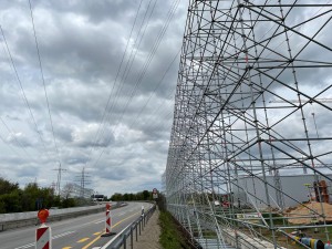 Eine Straßenbaustelle mit einem hohen, komplexen Gerüst aus Metallstangen entlang der rechten Seite einer Straße. Die Straße ist durch Leitplanken und rot-weiße Baustellenbaken mit Warnlichtern abgesichert. Darüber verlaufen zahlreiche Hochspannungsleitungen. Im Hintergrund sind Kräne und Bauarbeiten sichtbar. Der Himmel ist bewölkt.