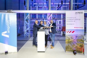 Das Bild zeigt vier Männer in Business-Kleidung, die gemeinsam symbolisch einen großen Schalter oder Hebel betätigen. Sie stehen in einer modernen Industriehalle mit technischen Anlagen und lila-blauer Beleuchtung. Links und rechts befinden sich zwei große Banner: eines mit dem Logo von Amprion und der Aufschrift „verbündet“, das andere mit dem Hitachi Energy-Logo und dem Slogan „Advancing a sustainable energy future for all“. Die Szenerie deutet auf die Inbetriebnahme oder Einweihung einer neuen Energieanlage hin. Die Männer lächeln und wirken feierlich.