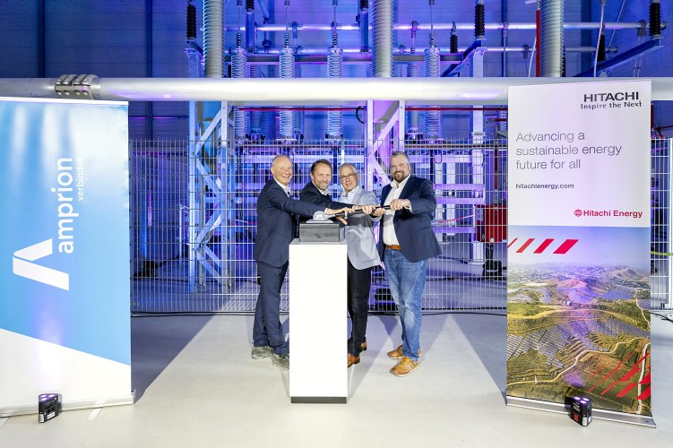 Das Bild zeigt vier Männer in Business-Kleidung, die gemeinsam symbolisch einen großen Schalter oder Hebel betätigen. Sie stehen in einer modernen Industriehalle mit technischen Anlagen und lila-blauer Beleuchtung. Links und rechts befinden sich zwei große Banner: eines mit dem Logo von Amprion und der Aufschrift „verbündet“, das andere mit dem Hitachi Energy-Logo und dem Slogan „Advancing a sustainable energy future for all“. Die Szenerie deutet auf die Inbetriebnahme oder Einweihung einer neuen Energieanlage hin. Die Männer lächeln und wirken feierlich.