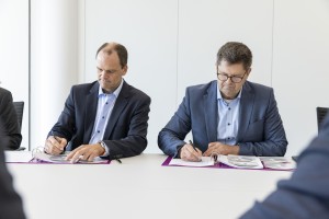 Das Bild zeigt zwei Männer in Business-Kleidung, die an einem weißen Tisch sitzen und Dokumente in lila Ordnern unterschreiben. Beide tragen dunkle Sakkos mit hellblauen Hemden. Der Mann links hält einen Stift und zeigt auf eine Seite, während der Mann rechts mit Brille konzentriert seine Unterschrift setzt. Die Szenerie wirkt förmlich und professionell, möglicherweise handelt es sich um eine Vertragsunterzeichnung oder eine offizielle Vereinbarung. Im Hintergrund ist eine schlichte, helle Wand zu sehen, und im Vordergrund sind leicht verschwommene Personen angedeutet, die an dem Meeting teilnehmen.