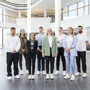 Das Bild zeigt eine Gruppe junger Menschen in einem modernen Bürogebäude, vermutlich Auszubildende oder neue Mitarbeiter.
Details:
Acht Personen, gemischt männlich und weiblich, stehen in einer Reihe.
Sie tragen eine Mischung aus formeller und lockerer Kleidung, meist Blazer oder Strickpullover.
Alle haben freundliche, selbstbewusste Ausdrücke.
Die Kulisse ist ein modernes Bürofoyer mit großen Glasfenstern, weißen Säulen und geschwungenen LED-Leuchten.
Dieses Bild vermittelt eine positive und moderne Unternehmenskultur, die Nachwuchstalente fördert.