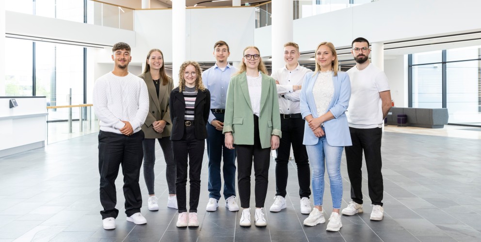 Das Bild zeigt eine Gruppe junger Menschen in einem modernen Bürogebäude, vermutlich Auszubildende oder neue Mitarbeiter.
Details:
Acht Personen, gemischt männlich und weiblich, stehen in einer Reihe.
Sie tragen eine Mischung aus formeller und lockerer Kleidung, meist Blazer oder Strickpullover.
Alle haben freundliche, selbstbewusste Ausdrücke.
Die Kulisse ist ein modernes Bürofoyer mit großen Glasfenstern, weißen Säulen und geschwungenen LED-Leuchten.
Dieses Bild vermittelt eine positive und moderne Unternehmenskultur, die Nachwuchstalente fördert.