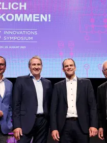Dieses Bild zeigt vier Männer auf der Bühne des Innovationssymposiums 2023 von Amprion.
Details:
Im Hintergrund befindet sich die große digitale Leinwand mit der Begrüßungsbotschaft des Events, ähnlich wie im vorherigen Bild.
Die vier Männer stehen entspannt und lächeln in die Kamera.
Einer der Männer trägt ein auffälliges hellblaues Sakko, was ihn visuell hervorhebt.
Die Bühne und das Event-Branding sind professionell gestaltet, mit dunkler Beleuchtung und lila Akzenten.
