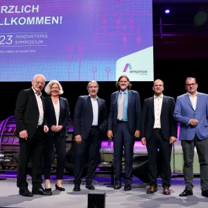 Das Bild zeigt sechs Personen auf einer Bühne beim Innovationssymposium 2023 von Amprion, das am 25. August 2023 in der Zeche Zollverein, Essen, stattfand.
Details:
Hinter der Gruppe befindet sich eine große digitale Leinwand mit der Aufschrift:
 "HERZLICH WILLKOMMEN! – 2023 INNOVATIONS SYMPOSIUM" sowie das Amprion-Logo.
Die Bühne ist modern beleuchtet, mit violettem Licht und historischen Maschinen im Hintergrund.
Auf der rechten Seite steht ein Rednerpult mit der Aufschrift des Events.
Die Gruppe besteht aus fünf Männern und einer Frau, alle in Business-Kleidung, lächelnd und in aufrechter Haltung.