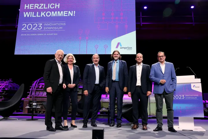 Das Bild zeigt sechs Personen auf einer Bühne beim Innovationssymposium 2023 von Amprion, das am 25. August 2023 in der Zeche Zollverein, Essen, stattfand.
Details:
Hinter der Gruppe befindet sich eine große digitale Leinwand mit der Aufschrift:
 "HERZLICH WILLKOMMEN! – 2023 INNOVATIONS SYMPOSIUM" sowie das Amprion-Logo.
Die Bühne ist modern beleuchtet, mit violettem Licht und historischen Maschinen im Hintergrund.
Auf der rechten Seite steht ein Rednerpult mit der Aufschrift des Events.
Die Gruppe besteht aus fünf Männern und einer Frau, alle in Business-Kleidung, lächelnd und in aufrechter Haltung.