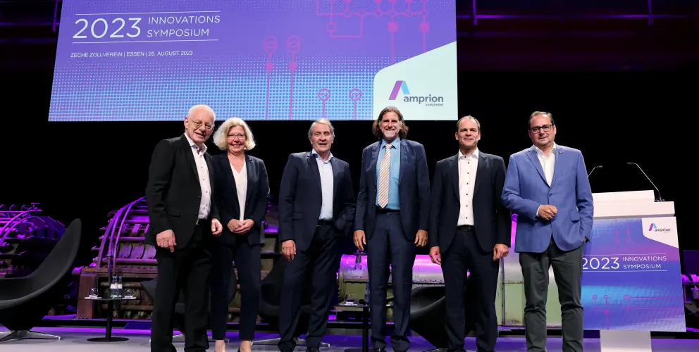 Das Bild zeigt sechs Personen auf einer Bühne beim Innovationssymposium 2023 von Amprion, das am 25. August 2023 in der Zeche Zollverein, Essen, stattfand.
Details:
Hinter der Gruppe befindet sich eine große digitale Leinwand mit der Aufschrift:
 "HERZLICH WILLKOMMEN! – 2023 INNOVATIONS SYMPOSIUM" sowie das Amprion-Logo.
Die Bühne ist modern beleuchtet, mit violettem Licht und historischen Maschinen im Hintergrund.
Auf der rechten Seite steht ein Rednerpult mit der Aufschrift des Events.
Die Gruppe besteht aus fünf Männern und einer Frau, alle in Business-Kleidung, lächelnd und in aufrechter Haltung.