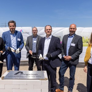 Das Bild zeigt sechs Personen in Business-Kleidung bei der feierlichen Grundsteinlegung einer Amprion-Konverterstation. Sie stehen hinter einem symbolischen Grundstein aus weißen Ziegeln mit einer Metallplatte, auf der die Inschrift „Amprion – Konverterstation Heidestück-Osterath – Grundsteinlegung 31. Mai 2023“ zu lesen ist.
Die Personen tragen teilweise Arbeitshandschuhe und halten Maurerkellen mit etwas Mörtel, was den symbolischen Charakter der Veranstaltung unterstreicht. Im Hintergrund sind ein Banner mit dem Amprion-Logo sowie eine Baustelle mit einem Bauarbeiter in orangefarbener Schutzkleidung zu sehen. Der Himmel ist klar und blau, was die festliche Atmosphäre verstärkt.
