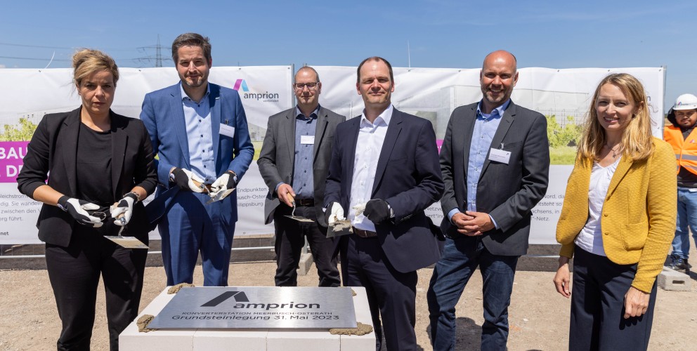 Das Bild zeigt sechs Personen in Business-Kleidung bei der feierlichen Grundsteinlegung einer Amprion-Konverterstation. Sie stehen hinter einem symbolischen Grundstein aus weißen Ziegeln mit einer Metallplatte, auf der die Inschrift „Amprion – Konverterstation Heidestück-Osterath – Grundsteinlegung 31. Mai 2023“ zu lesen ist.
Die Personen tragen teilweise Arbeitshandschuhe und halten Maurerkellen mit etwas Mörtel, was den symbolischen Charakter der Veranstaltung unterstreicht. Im Hintergrund sind ein Banner mit dem Amprion-Logo sowie eine Baustelle mit einem Bauarbeiter in orangefarbener Schutzkleidung zu sehen. Der Himmel ist klar und blau, was die festliche Atmosphäre verstärkt.
