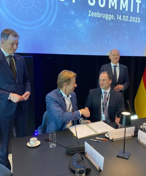 Das Bild zeigt eine feierliche Unterzeichnungszeremonie beim North Sea Summit am 14. Februar 2023 in Zeebrugge. Zwei Männer sitzen an einem Konferenztisch und schütteln sich die Hand, während sie ein Abkommen unterzeichnen. Hinter ihnen stehen zwei weitere Männer und beobachten die Szene mit freundlichen Ausdrücken.
Der Konferenztisch ist mit Mikrofonen, Dokumenten, Namensschildern, Wasser- und Kaffeegläsern sowie kleinen Tischlampen ausgestattet. Im Hintergrund sind die Flaggen von Deutschland, Belgien und der Europäischen Union zu sehen. Die Atmosphäre wirkt professionell und diplomatisch.