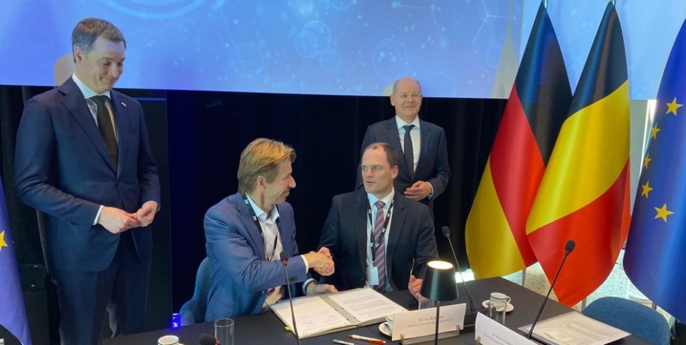 Das Bild zeigt eine feierliche Unterzeichnungszeremonie beim North Sea Summit am 14. Februar 2023 in Zeebrugge. Zwei Männer sitzen an einem Konferenztisch und schütteln sich die Hand, während sie ein Abkommen unterzeichnen. Hinter ihnen stehen zwei weitere Männer und beobachten die Szene mit freundlichen Ausdrücken.
Der Konferenztisch ist mit Mikrofonen, Dokumenten, Namensschildern, Wasser- und Kaffeegläsern sowie kleinen Tischlampen ausgestattet. Im Hintergrund sind die Flaggen von Deutschland, Belgien und der Europäischen Union zu sehen. Die Atmosphäre wirkt professionell und diplomatisch.