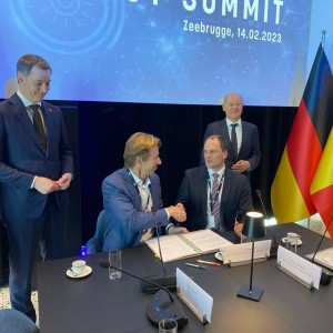 Das Bild zeigt eine feierliche Unterzeichnungszeremonie beim North Sea Summit am 14. Februar 2023 in Zeebrugge. Zwei Männer sitzen an einem Konferenztisch und schütteln sich die Hand, während sie ein Abkommen unterzeichnen. Hinter ihnen stehen zwei weitere Männer und beobachten die Szene mit freundlichen Ausdrücken.
Der Konferenztisch ist mit Mikrofonen, Dokumenten, Namensschildern, Wasser- und Kaffeegläsern sowie kleinen Tischlampen ausgestattet. Im Hintergrund sind die Flaggen von Deutschland, Belgien und der Europäischen Union zu sehen. Die Atmosphäre wirkt professionell und diplomatisch.