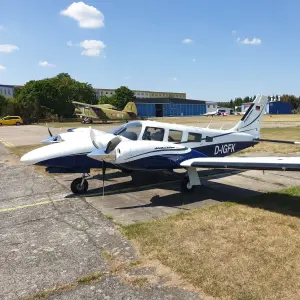 Im Vordergrund steht ein zweimotoriges Leichtflugzeug vom Typ Piper PA-34 Seneca mit der Kennung „D-IGFK“. Das Flugzeug ist weiß mit blauen Akzenten, hat zwei Propellertriebwerke und ist auf einer Betonfläche geparkt.
 Im Hintergrund ist ein weiteres, olivgrünes, älteres Flugzeug mit der Markierung „801“ zu sehen, das einem militärischen oder historischen Modell ähnelt. Dahinter befinden sich blaue Hangars, ein gelbes Auto und eine weitläufige Rasenfläche mit einer Landebahn. Der Himmel ist klar mit wenigen Wolken, und die Szenerie wirkt sommerlich.
