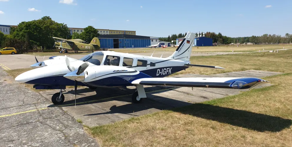 Im Vordergrund steht ein zweimotoriges Leichtflugzeug vom Typ Piper PA-34 Seneca mit der Kennung „D-IGFK“. Das Flugzeug ist weiß mit blauen Akzenten, hat zwei Propellertriebwerke und ist auf einer Betonfläche geparkt.
 Im Hintergrund ist ein weiteres, olivgrünes, älteres Flugzeug mit der Markierung „801“ zu sehen, das einem militärischen oder historischen Modell ähnelt. Dahinter befinden sich blaue Hangars, ein gelbes Auto und eine weitläufige Rasenfläche mit einer Landebahn. Der Himmel ist klar mit wenigen Wolken, und die Szenerie wirkt sommerlich.