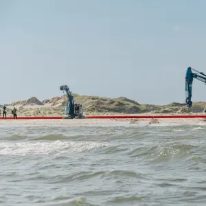 Das Bild zeigt eine Küstenszene mit zwei blauen Baggern auf einem Sandstrand, die an einer langen, roten Rohre arbeiten. Im Vordergrund ist das Meer mit kleinen Wellen zu sehen. Mehrere Personen in Sicherheitswesten und Schutzkleidung befinden sich in der Nähe der Maschinen und scheinen an der Installation oder Wartung der Barriere beteiligt zu sein. Im Hintergrund erstrecken sich sandige Dünen mit spärlicher Vegetation unter einem klaren blauen Himmel.