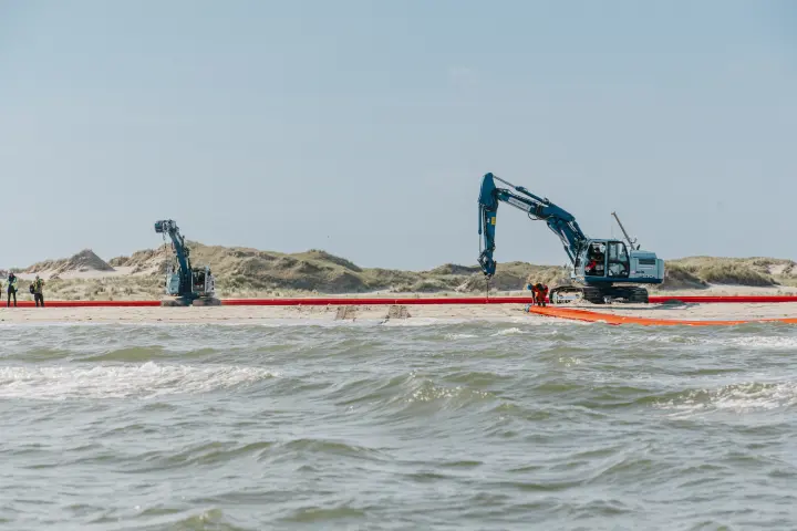 Das Bild zeigt eine Küstenszene mit zwei blauen Baggern auf einem Sandstrand, die an einer langen, roten Rohre arbeiten. Im Vordergrund ist das Meer mit kleinen Wellen zu sehen. Mehrere Personen in Sicherheitswesten und Schutzkleidung befinden sich in der Nähe der Maschinen und scheinen an der Installation oder Wartung der Barriere beteiligt zu sein. Im Hintergrund erstrecken sich sandige Dünen mit spärlicher Vegetation unter einem klaren blauen Himmel.