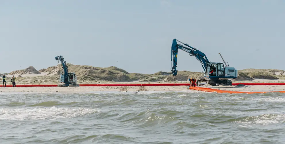 Das Bild zeigt eine Küstenszene mit zwei blauen Baggern auf einem Sandstrand, die an einer langen, roten Rohre arbeiten. Im Vordergrund ist das Meer mit kleinen Wellen zu sehen. Mehrere Personen in Sicherheitswesten und Schutzkleidung befinden sich in der Nähe der Maschinen und scheinen an der Installation oder Wartung der Barriere beteiligt zu sein. Im Hintergrund erstrecken sich sandige Dünen mit spärlicher Vegetation unter einem klaren blauen Himmel.