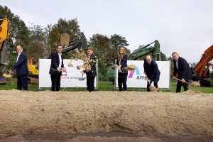 Das Bild zeigt sechs Personen in formeller Kleidung, die bei einem symbolischen Spatenstich Sand mit Schaufeln in die Luft werfen. Hinter ihnen stehen mehrere Bagger in verschiedenen Farben, und es sind Banner mit Logos und Schriftzügen zu sehen, darunter „amp“ und „GEMEINSAM für die Energiewende“. Der Hintergrund besteht aus grünen Bäumen und einer Wiese, was auf eine Baustelle in einem eher ländlichen oder naturnahen Gebiet hindeutet. Die Personen wirken gut gelaunt und engagiert.