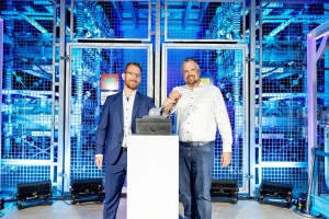 Das Bild zeigt zwei Männer, die gemeinsam einen symbolischen Knopf drücken. Der Mann links trägt einen dunkelblauen Anzug mit weißem Hemd und hat einen kurzen Bart sowie eine Brille. Der Mann rechts trägt ein helles Hemd mit hochgekrempelten Ärmeln und eine Jeans. Beide lächeln und schauen in die Kamera.
Der Hintergrund ist von einem industriellen oder technologischen Umfeld geprägt, mit blauen Gitterstrukturen und technischer Ausstattung. Die Szene ist hell beleuchtet, mit blauen und weißen Lichteffekten.