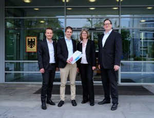 Vier Personen stehen vor dem Gebäude der Bundesnetzagentur, erkennbar am Wappen mit dem Bundesadler und der Aufschrift "Bundesnetzagentur" auf der Glasscheibe.
Details:
Die Gruppe trägt Business-Kleidung, zwei Männer und eine Frau in Anzügen.
Eine Person hält einen Stapel Dokumente mit einer Amprion-Mappe, was auf eine formelle Einreichung hindeutet.
Die Kulisse ist ein modernes Verwaltungsgebäude mit Glasfassade.