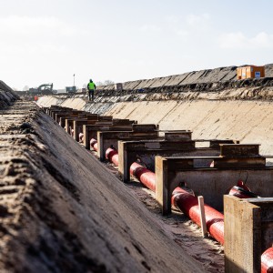 Das Bild zeigt eine Baustelle für eine Erdkabeltrasse aus einer bodennahen Perspektive.
Details zur Szene:
Im Vordergrund: Mehrere rote Kabel sind in einem ausgehobenen Graben verlegt und liegen auf stabilen Metallhalterungen. Diese Konstruktion sorgt für die korrekte Positionierung der Kabel.
Der Graben: Die Seiten des Grabens sind glatt und schräg abfallend, um ein Abrutschen der Erde zu verhindern.
Hintergrund:
Ein Bauarbeiter mit Warnweste und Schutzhelm geht entlang der Baustelle.
Ein Bagger ist im Einsatz, wahrscheinlich für Aushubarbeiten.
Ein orangefarbener Container steht am Rand der Baustelle, vermutlich als Baubüro oder Lager.
Aufgeschüttete Erdhügel sind zu sehen, was auf laufende Bauarbeiten hinweist.
Licht und Umgebung:
Der Himmel ist teilweise bewölkt, aber es scheint die Sonne, wodurch starke Kontraste entstehen.
Der Boden ist trocken und sandig