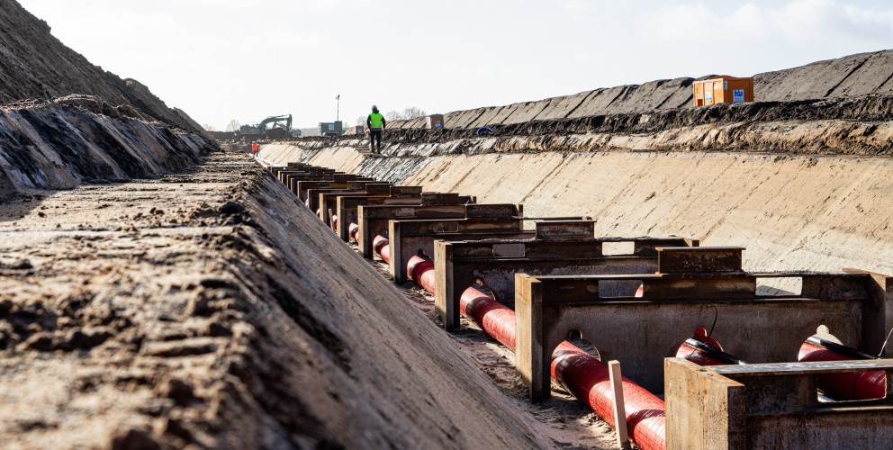 Das Bild zeigt eine Baustelle für eine Erdkabeltrasse aus einer bodennahen Perspektive.
Details zur Szene:
Im Vordergrund: Mehrere rote Kabel sind in einem ausgehobenen Graben verlegt und liegen auf stabilen Metallhalterungen. Diese Konstruktion sorgt für die korrekte Positionierung der Kabel.
Der Graben: Die Seiten des Grabens sind glatt und schräg abfallend, um ein Abrutschen der Erde zu verhindern.
Hintergrund:
Ein Bauarbeiter mit Warnweste und Schutzhelm geht entlang der Baustelle.
Ein Bagger ist im Einsatz, wahrscheinlich für Aushubarbeiten.
Ein orangefarbener Container steht am Rand der Baustelle, vermutlich als Baubüro oder Lager.
Aufgeschüttete Erdhügel sind zu sehen, was auf laufende Bauarbeiten hinweist.
Licht und Umgebung:
Der Himmel ist teilweise bewölkt, aber es scheint die Sonne, wodurch starke Kontraste entstehen.
Der Boden ist trocken und sandig