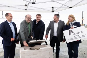 Das Bild zeigt fünf Personen bei einer Grundsteinlegung auf einer Baustelle. Sie stehen gemeinsam um einen Steinblock mit einer frisch aufgetragenen Schicht Mörtel. In der Mitte hält ein Mann mit Brille und grünem Hemd eine schwarze Kapsel, die vermutlich in den Grundstein eingebettet wird. Die anderen Personen, bestehend aus drei Männern und einer Frau, lächeln und schauen in die Kamera.
Die Frau rechts hält ein Schild mit dem Logo von "Amprion" und der Aufschrift:
 „A-Nord Konverterstation Emden-Ost Grundsteinlegung 12. Juni 2024.“
Der Hintergrund zeigt ein großes Zelt mit Firmenlogos und Baustellenelementen, was auf eine feierliche Veranstaltung in einem professionellen Rahmen hinweist. Alle Personen tragen Business-Kleidung und vermitteln einen offiziellen und positiven Eindruck.