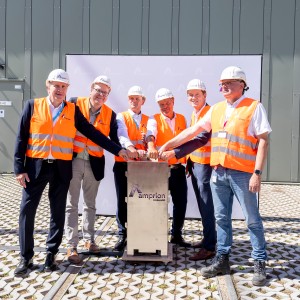 Das Bild zeigt sechs Männer bei einer feierlichen Projektveranstaltung. Sie tragen orangene Warnwesten und weiße Helme, was auf eine Baustelle oder ein technisches Umfeld hinweist. Alle Männer stehen um einen Schaltkasten mit dem Amprion-Logo und legen gemeinsam die Hände auf einen symbolischen Knopf oder Schalter.
Im Hintergrund befindet sich eine große industrielle Anlage oder Halle mit einer Metallwand. Der Boden ist gepflastert, und eine weiße Leinwand mit Firmenlogos steht hinter der Gruppe. Die Männer lächeln und schauen in die Kamera, was die feierliche und positive Atmosphäre der Veranstaltung betont.
