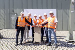 Das Bild zeigt sechs Männer bei einer feierlichen Projektveranstaltung. Sie tragen orangene Warnwesten und weiße Helme, was auf eine Baustelle oder ein technisches Umfeld hinweist. Alle Männer stehen um einen Schaltkasten mit dem Amprion-Logo und legen gemeinsam die Hände auf einen symbolischen Knopf oder Schalter.
Im Hintergrund befindet sich eine große industrielle Anlage oder Halle mit einer Metallwand. Der Boden ist gepflastert, und eine weiße Leinwand mit Firmenlogos steht hinter der Gruppe. Die Männer lächeln und schauen in die Kamera, was die feierliche und positive Atmosphäre der Veranstaltung betont.
