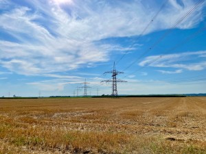 Das Bild zeigt eine weite, abgeerntete Feldlandschaft unter einem teils bewölkten blauen Himmel. Mehrere hohe Strommasten aus Metall mit daran verlaufenden Hochspannungsleitungen ziehen sich durch die Szenerie. Im Hintergrund sind grüne Felder und eine leicht hügelige Landschaft zu sehen. Die Sonne scheint, und das Licht erzeugt weiche Schatten auf dem trockenen Boden.