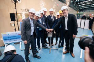 Das Bild zeigt eine Gruppe von acht Männern in einem industriellen Umfeld, die gemeinsam einen symbolischen Knopf drücken. Sie tragen Anzüge und Schutzhelme, einige mit Firmenlogos. Im Hintergrund sprühen Funken, und es gibt Schilder mit der Aufschrift "Brennstart DolWin delta | S.727" sowie das Datum 04. Juli 2024. Einige Personen im Hintergrund tragen ebenfalls Helme und Schutzkleidung, während eine Kamera im Vordergrund die Szene festhält. Die feierliche Atmosphäre deutet auf den Start eines wichtigen Projekts im Energiesektor hin, wahrscheinlich im Bereich Offshore-Windenergie.