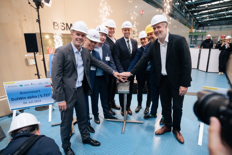 Das Bild zeigt eine Gruppe von acht Männern in einem industriellen Umfeld, die gemeinsam einen symbolischen Knopf drücken. Sie tragen Anzüge und Schutzhelme, einige mit Firmenlogos. Im Hintergrund sprühen Funken, und es gibt Schilder mit der Aufschrift "Brennstart DolWin delta | S.727" sowie das Datum 04. Juli 2024. Einige Personen im Hintergrund tragen ebenfalls Helme und Schutzkleidung, während eine Kamera im Vordergrund die Szene festhält. Die feierliche Atmosphäre deutet auf den Start eines wichtigen Projekts im Energiesektor hin, wahrscheinlich im Bereich Offshore-Windenergie.