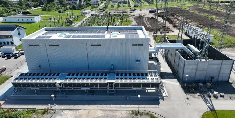 Im Mittelpunkt steht ein großes, rechteckiges Industriegebäude mit einer metallverkleideten Fassade und mehreren Lüftungsgittern. Auf dem Dach sind Solarpanels zu sehen.
Vor dem Gebäude befindet sich eine lange Reihe von großen Kühlanlagen mit zahlreichen Ventilatoren. Rechts daneben verlaufen mehrere große Stromleitungen, die über hohe Gittermasten geführt werden. Im Hintergrund sind weitere Infrastrukturelemente einer Umspannanlage sichtbar, darunter Schaltanlagen, Freilufttransformatoren und mehrere kleine Betriebsgebäude.
Die Umgebung besteht aus grünen Wiesen, Straßen und einigen parkenden Fahrzeugen.
