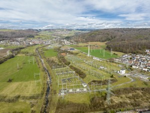 Das Bild zeigt eine große Umspannanlage in einer ländlichen Umgebung aus der Luftperspektive. Die Anlage besteht aus einem weitläufigen Netzwerk von Strommasten, Hochspannungsleitungen und Schaltanlagen, die über eine grüne Wiesenlandschaft verteilt sind.
Ein kleiner Fluss schlängelt sich durch das Gelände, und mehrere hohe Stromleitungen führen aus der Umspannanlage heraus. Im Hintergrund sind Wohnsiedlungen mit weißen Häusern und roten Dächern zu sehen, eingebettet in eine hügelige Landschaft mit Feldern, Wäldern und weiteren Strommasten.
Der Himmel ist leicht bewölkt, und die Szene vermittelt den Eindruck einer gut vernetzten Infrastruktur zur Energieübertragung in einer naturnahen Umgebung.