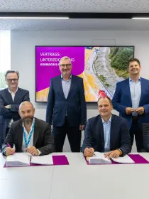 Das Bild zeigt eine feierliche Vertragsunterzeichnung mit sieben Männern in Business-Kleidung. Vier von ihnen sitzen an einem Tisch und unterschreiben Dokumente, während drei weitere hinter ihnen stehen und lächeln. Jeder der Unterzeichner hat einen farbigen Stift in der Hand und vor sich große Ordner mit Dokumenten auf lilafarbenen Mappen.
Im Hintergrund ist ein Bildschirm mit der Aufschrift 