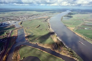 
Das Luftbild zeigt eine weitläufige Flusslandschaft mit einem großen, mäandrierenden Strom, kleineren Nebenarmen und grünen Wiesen. Ein Frachtschiff fährt auf dem breiten Hauptfluss. Links im Bild befindet sich eine Siedlung mit zahlreichen Häusern und Gewerbegebäuden, während auf der rechten Seite landwirtschaftlich genutzte Flächen mit schmalen Entwässerungskanälen zu sehen sind.
Die Landschaft ist geprägt von Deichen, Weideflächen mit Schafen und mehreren Wasserwegen. Im Hintergrund erstrecken sich große Felder und Windkraftanlagen entlang des Horizonts. Der Himmel ist teilweise bewölkt, und die Wolkenschatten werfen dunkle Muster auf die Landschaft.
