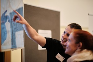 Das Bild zeigt eine Informationsveranstaltung oder einen Bürgerdialog, bei dem ein Mitarbeiter einem Besucher eine digitale Karte oder ein Planungsdokument erklärt.
Szene:
Ein Mann mit Namensschild "Tom Kaufner" in schwarzem Polohemd zeigt auf einen großen Touchscreen.
Eine Frau mit rotem Haar und Schal betrachtet die Anzeige interessiert.
Im Hintergrund sind Infotafeln zu sehen, die auf eine Präsentationsveranstaltung hindeuten.
Das Bild vermittelt eine offene und dialogorientierte Atmosphäre, die auf Bürgerbeteiligung oder öffentliche Information zu Infrastrukturprojekten schließen lässt.
