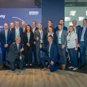 Das Bild zeigt eine große Gruppe von Geschäftsleuten bei einer feierlichen Unterzeichnungszeremonie auf einer Messe oder Konferenz.
Wichtige Elemente:
Im Hintergrund ist eine digitale Leinwand mit der Aufschrift:
 "Signature Ceremony – Letter of Intent"
 "Delivery of a F-gas free 420kV High Voltage Circuit Breaker",
 with Amprion GmbH und Siemens Energy-Logo.
Die Gruppe besteht aus Männern und Frauen in Business-Kleidung.
Einige Personen geben Daumen-hoch-Gesten, was auf eine erfolgreiche Vereinbarung hindeutet.
Die Umgebung wirkt modern, mit beleuchteten Wänden und Konferenztechnik.