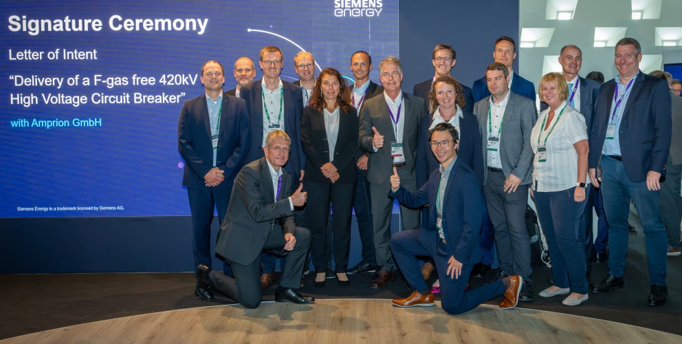 Das Bild zeigt eine große Gruppe von Geschäftsleuten bei einer feierlichen Unterzeichnungszeremonie auf einer Messe oder Konferenz.
Wichtige Elemente:
Im Hintergrund ist eine digitale Leinwand mit der Aufschrift:
 "Signature Ceremony – Letter of Intent"
 "Delivery of a F-gas free 420kV High Voltage Circuit Breaker",
 with Amprion GmbH und Siemens Energy-Logo.
Die Gruppe besteht aus Männern und Frauen in Business-Kleidung.
Einige Personen geben Daumen-hoch-Gesten, was auf eine erfolgreiche Vereinbarung hindeutet.
Die Umgebung wirkt modern, mit beleuchteten Wänden und Konferenztechnik.