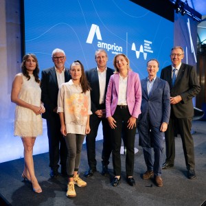 Das Bild zeigt eine Gruppe von sieben Personen auf einer Bühne vor einem großen blauen Bildschirm mit dem Amprion-Logo und der Aufschrift „verbindet“. Die Personen sind formell oder smart-casual gekleidet, einige tragen Anzüge, während andere modern-legere Outfits gewählt haben. Die Gruppe besteht aus Männern und Frauen, die entspannt und freundlich in die Kamera lächeln. Die Szenerie deutet auf eine professionelle Veranstaltung oder Konferenz hin, möglicherweise im Bereich Energie oder Infrastruktur. Die Bühne ist modern gestaltet, mit stilvoller Beleuchtung und einem technisch ausgestatteten Hintergrund.