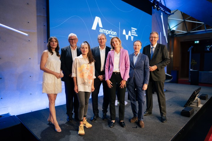 Das Bild zeigt eine Gruppe von sieben Personen auf einer Bühne vor einem großen blauen Bildschirm mit dem Amprion-Logo und der Aufschrift „verbindet“. Die Personen sind formell oder smart-casual gekleidet, einige tragen Anzüge, während andere modern-legere Outfits gewählt haben. Die Gruppe besteht aus Männern und Frauen, die entspannt und freundlich in die Kamera lächeln. Die Szenerie deutet auf eine professionelle Veranstaltung oder Konferenz hin, möglicherweise im Bereich Energie oder Infrastruktur. Die Bühne ist modern gestaltet, mit stilvoller Beleuchtung und einem technisch ausgestatteten Hintergrund.