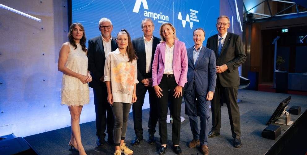 Das Bild zeigt eine Gruppe von sieben Personen auf einer Bühne vor einem großen blauen Bildschirm mit dem Amprion-Logo und der Aufschrift „verbindet“. Die Personen sind formell oder smart-casual gekleidet, einige tragen Anzüge, während andere modern-legere Outfits gewählt haben. Die Gruppe besteht aus Männern und Frauen, die entspannt und freundlich in die Kamera lächeln. Die Szenerie deutet auf eine professionelle Veranstaltung oder Konferenz hin, möglicherweise im Bereich Energie oder Infrastruktur. Die Bühne ist modern gestaltet, mit stilvoller Beleuchtung und einem technisch ausgestatteten Hintergrund.